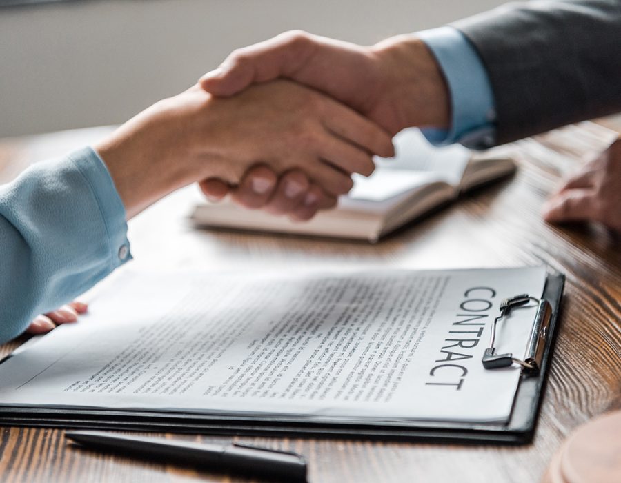 close-up view of clipboard with contract and lawyer with client close-up view of clipboard with contract and lawyer with client shaking hands behind